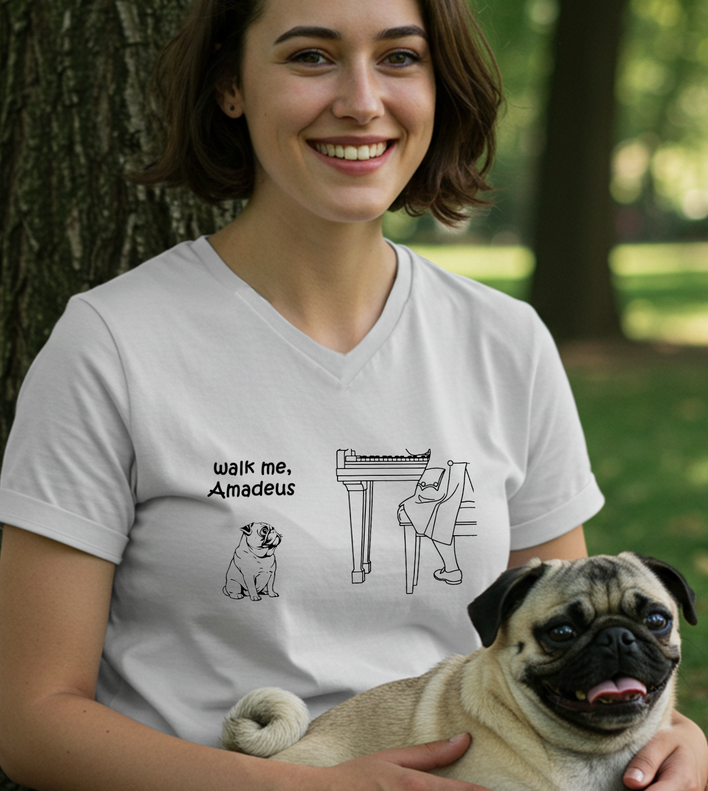 A woman wearing a white graphic t-shirt with a pug, sitting outdoors in a park next to a tree. The t-shirt features a black line art graphic of a pug and Mozart at a piano with the text 'walk me, Amadeus'.