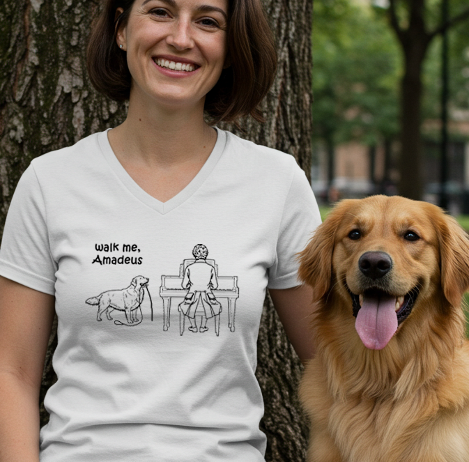 A woman wearing a white graphic t-shirt stands outdoors next to a golden retriever dog in front of a tree. The t-shirt features a black line art illustration of Mozart playing a piano with a Golden Retriever standing beside him, accompanied by the text 'walk me, Amadeus'.