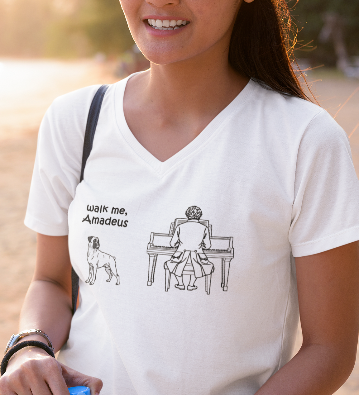 A woman wearing a white V-neck t-shirt with a black graphic print that reads 'walk me, Amadeus' next to an illustration of a Boxer standing beside Mozart at the piano. 