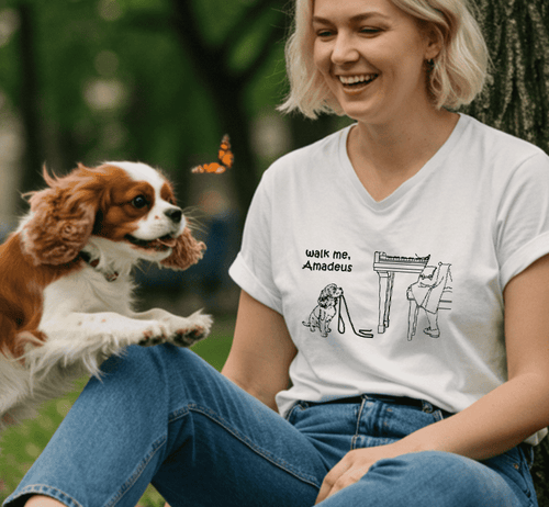 A woman sitting outdoors next to a tree wears a white graphic T-shirt with a black line drawing and the text 'walk me, Amadeu