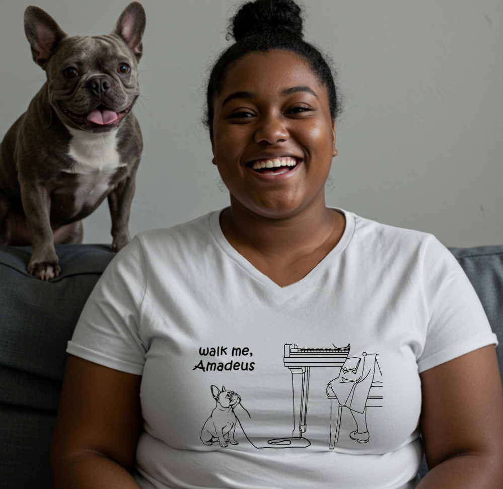 A smiling woman wearing a white graphic t-shirt with a black line drawing of a dog and the text 'walk me, Amadeus', with a grey French Bulldog sitting on the couch behind her.