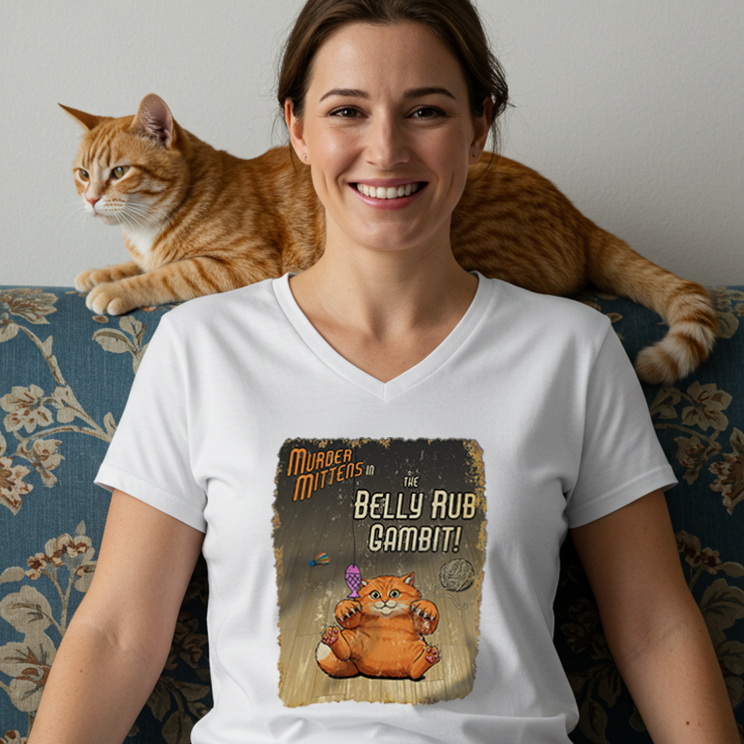 A woman sitting on a blue floral couch wearing a white V-neck t-shirt with a graphic that reads, 'Murder Mittens in the Belly Rub Gambit!'. An orange tabby cat is lying on the back of the couch behind her.