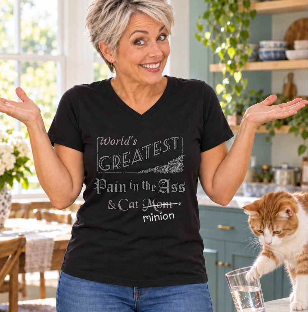 Woman shrugging in a kitchen beside a cat tipping a glass, wearing a black V-neck T-shirt reading “World’s Greatest Pain in the Ass & Cat Mom.” “Mom” is crossed out, with “minion” handwritten beneath it and surrounded by cat paw prints.