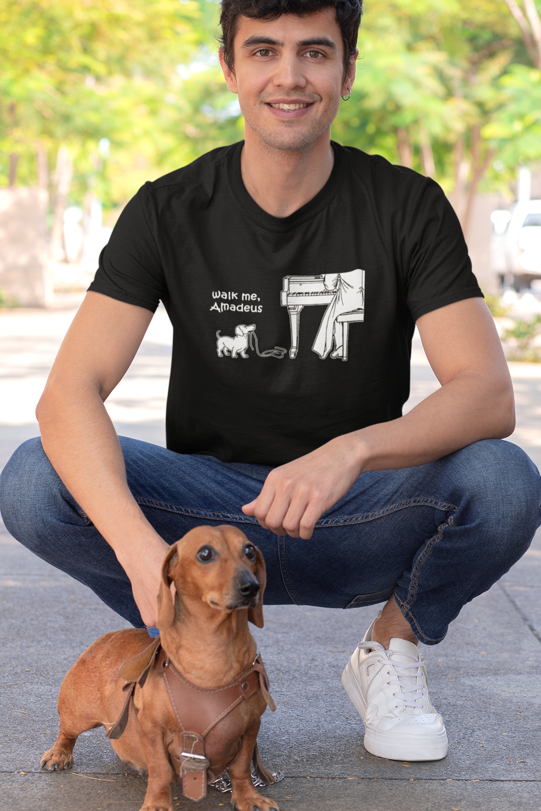 A man crouching outdoors on a sidewalk next to a dachshund wearing a black graphic t-shirt with a white print that reads ' Walk me, Amadeus'.