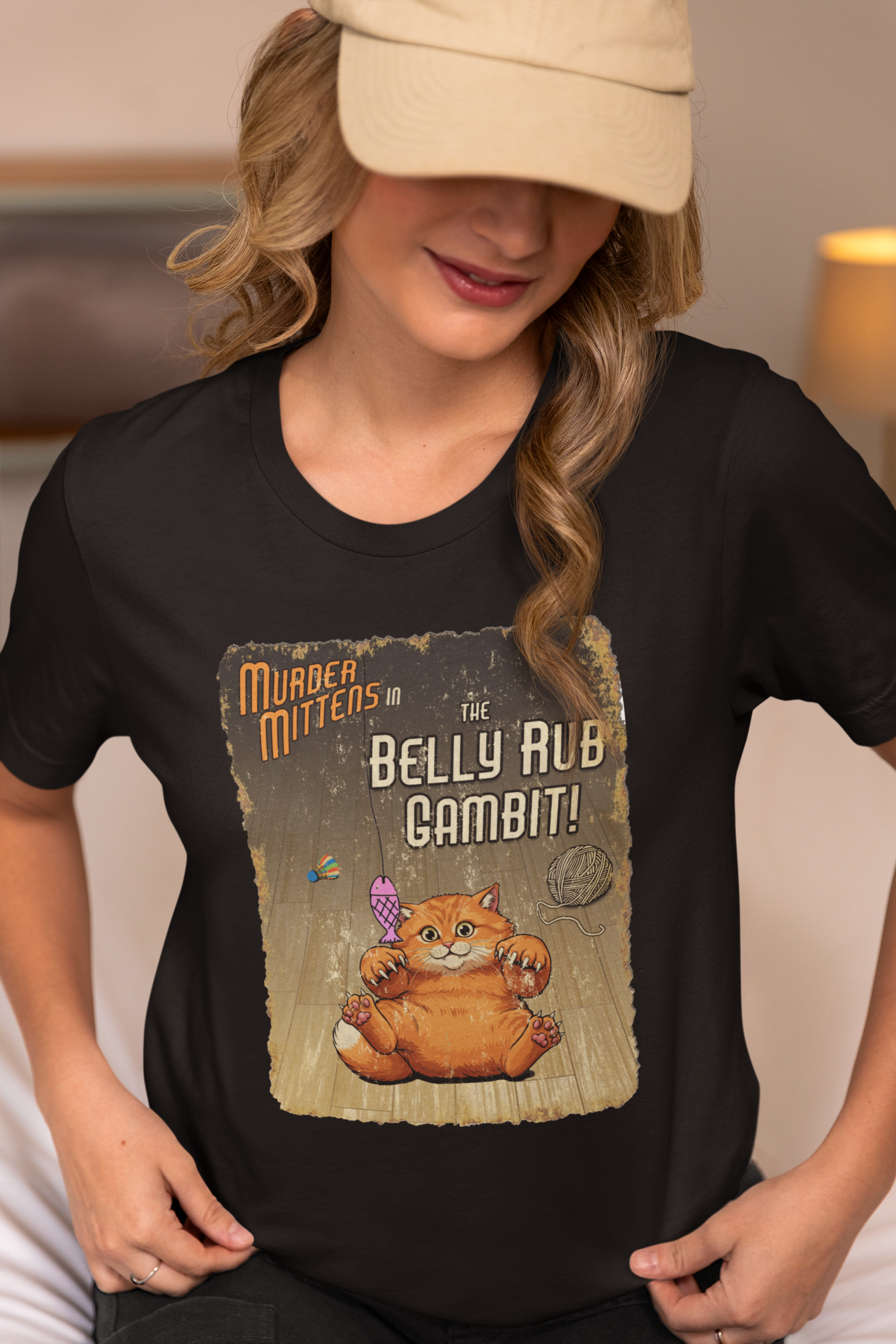 A woman wearing a black graphic t-shirt with a vintage-style print of an orange cat presenting his belly for a scratch, captioned 'Murder Mittens in the Belly Rub Gambit!'. 