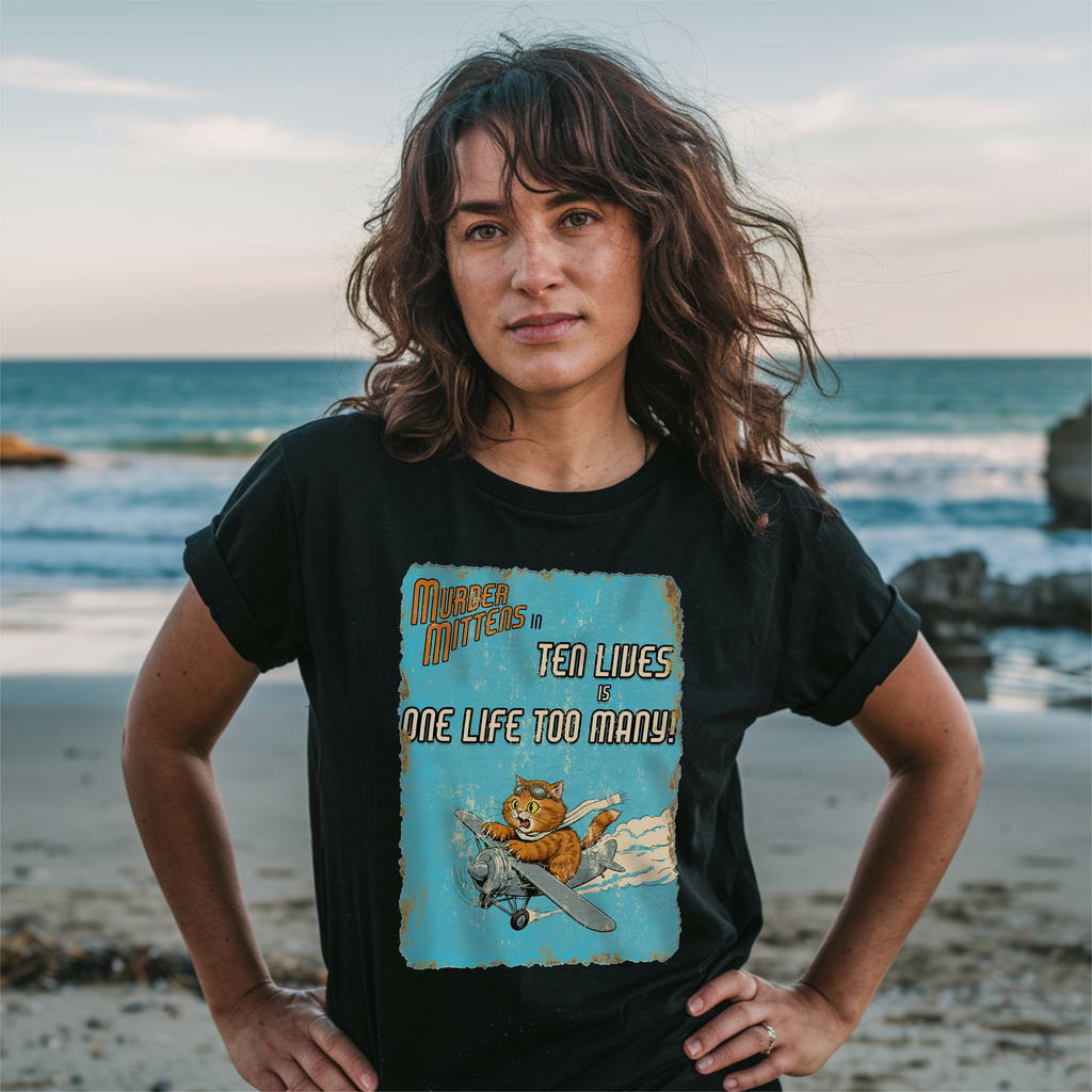A woman stands on a beach wearing a black graphic t-shirt. The shirt features a distressed light blue graphic with an orange cat flying a plane and the text 'Murder Mittens in TEN LIVES is ONE LIFE TOO MANY!'