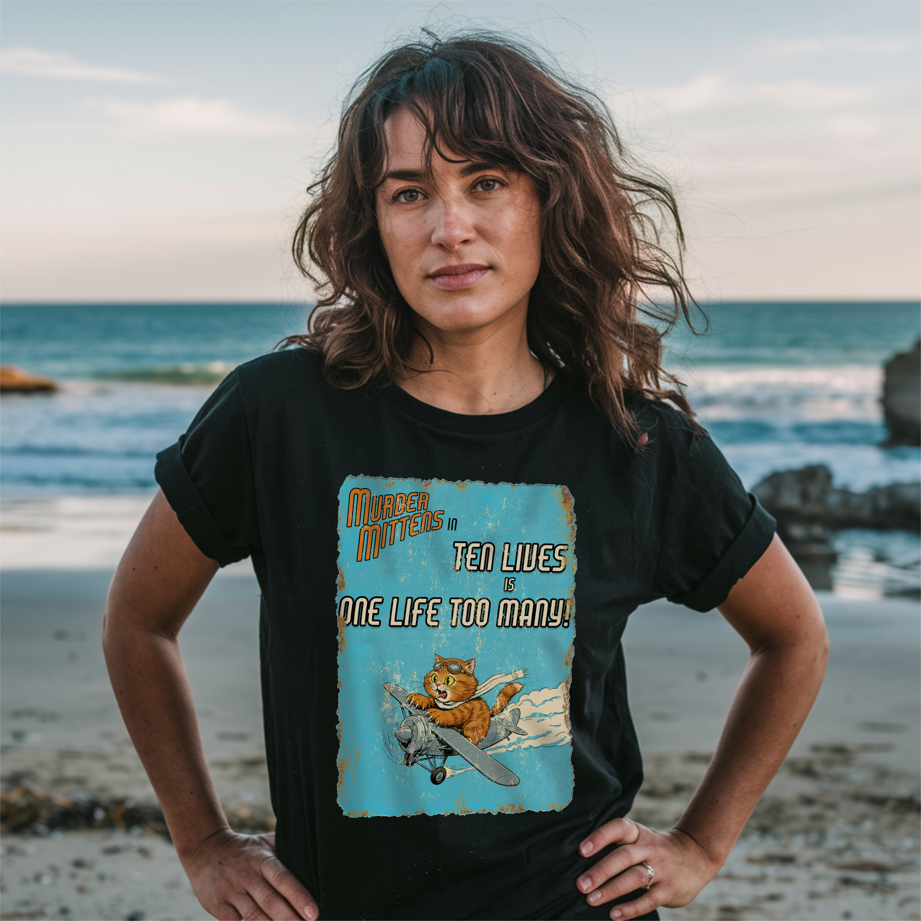 A woman stands on a beach wearing a black graphic t-shirt. The shirt features a distressed light blue graphic with an orange cat flying a plane and the text 'Murder Mittens in TEN LIVES is ONE LIFE TOO MANY!'