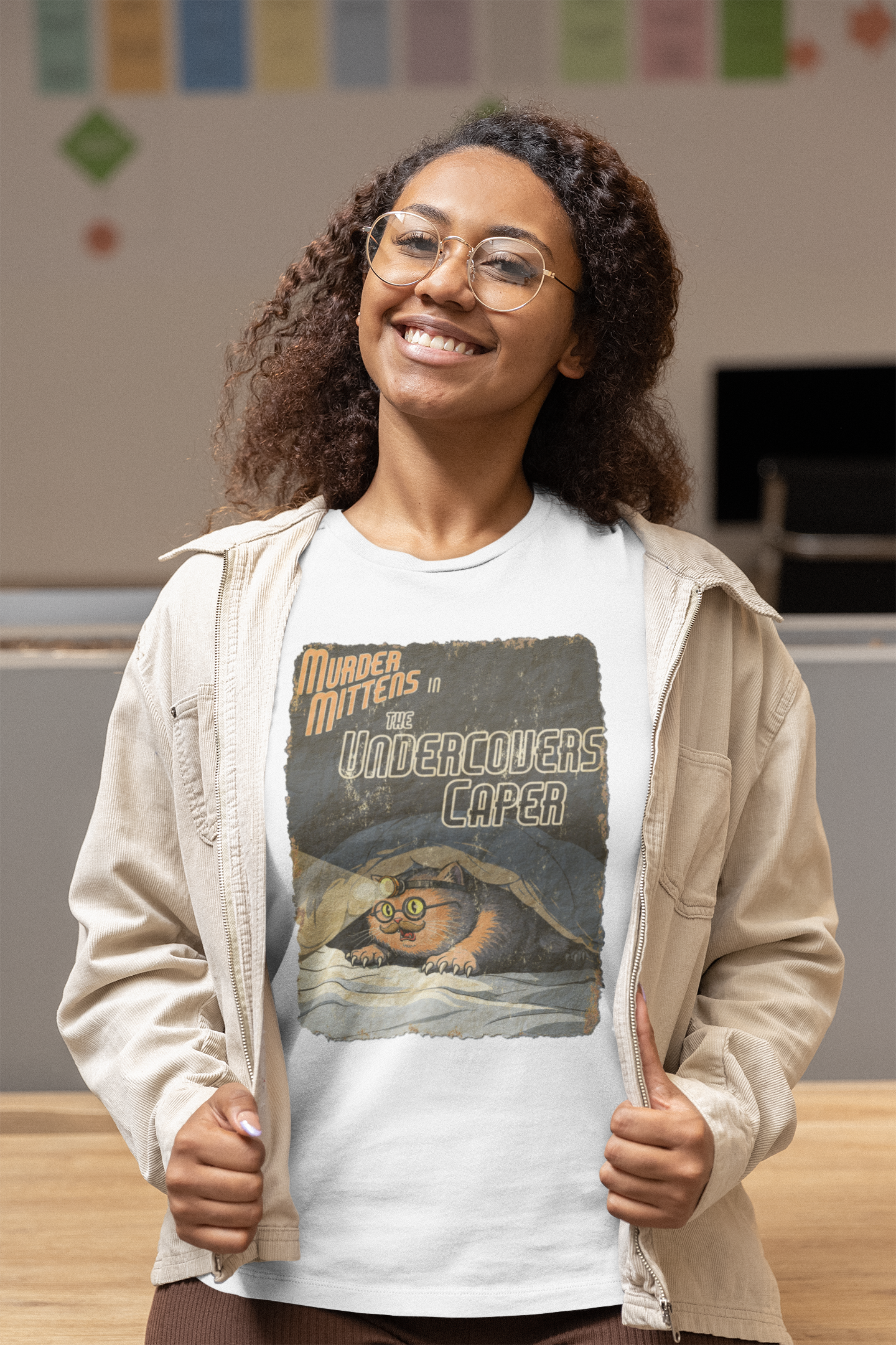 A smiling woman with curly hair and glasses wears a white graphic T-shirt under an open beige zip jacket in an indoor room setting. The T-shirt features a vintage-style illustrated print with the text 'Murder Mittens in the Undercovers Caper