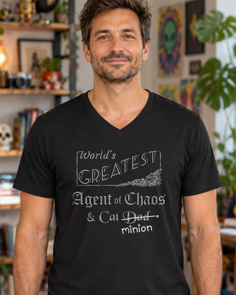 Man standing in an eclectic room, wearing a black V-neck t-shirt that reads, “World’s Greatest Agent of Chaos & Cat Dad” with “Dad” crossed out and “minion” handwritten below with paw prints.