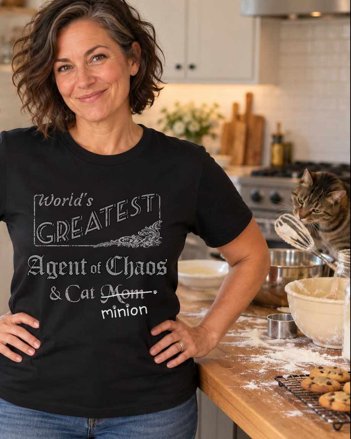 Woman in a kitchen beside baking supplies and a cat, wearing a black crew-neck t-shirt that reads, “World’s Greatest Agent of Chaos & Cat Mom” with “Mom” crossed out and “minion” handwritten below with paw prints.