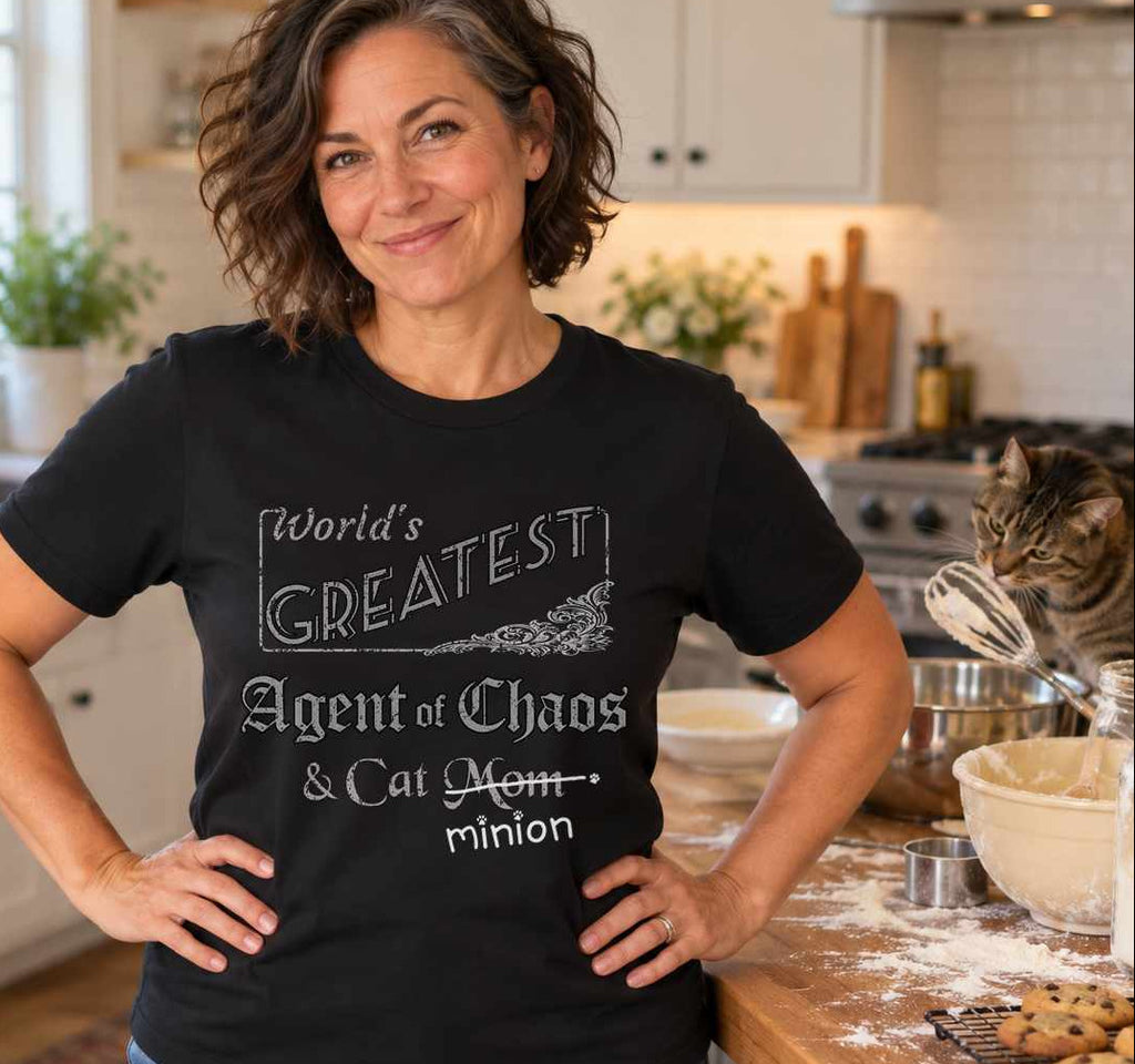 Woman in a kitchen beside baking supplies and a cat, wearing a black crew-neck t-shirt that reads, “World’s Greatest Agent of Chaos & Cat Mom” with “Mom” crossed out and “minion” handwritten below with paw prints.