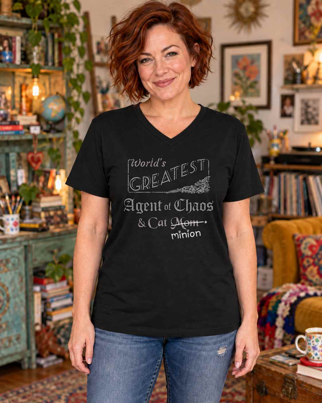 Woman standing in an eclectic room, wearing a black V-neck t-shirt that reads, “World’s Greatest Agent of Chaos & Cat Mom” with “Mom” crossed out and “minion” handwritten below with paw prints.