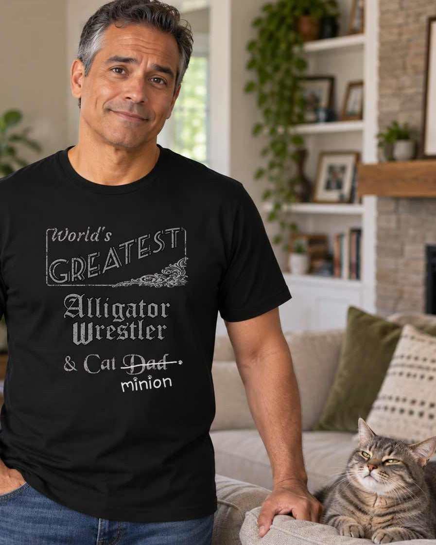 Man standing beside a cat on an armchair, wearing a black crew-neck t-shirt that reads, “World’s Greatest Alligator Wrestler & Cat Dad” with “Dad” crossed out and “minion” handwritten below with paw prints.