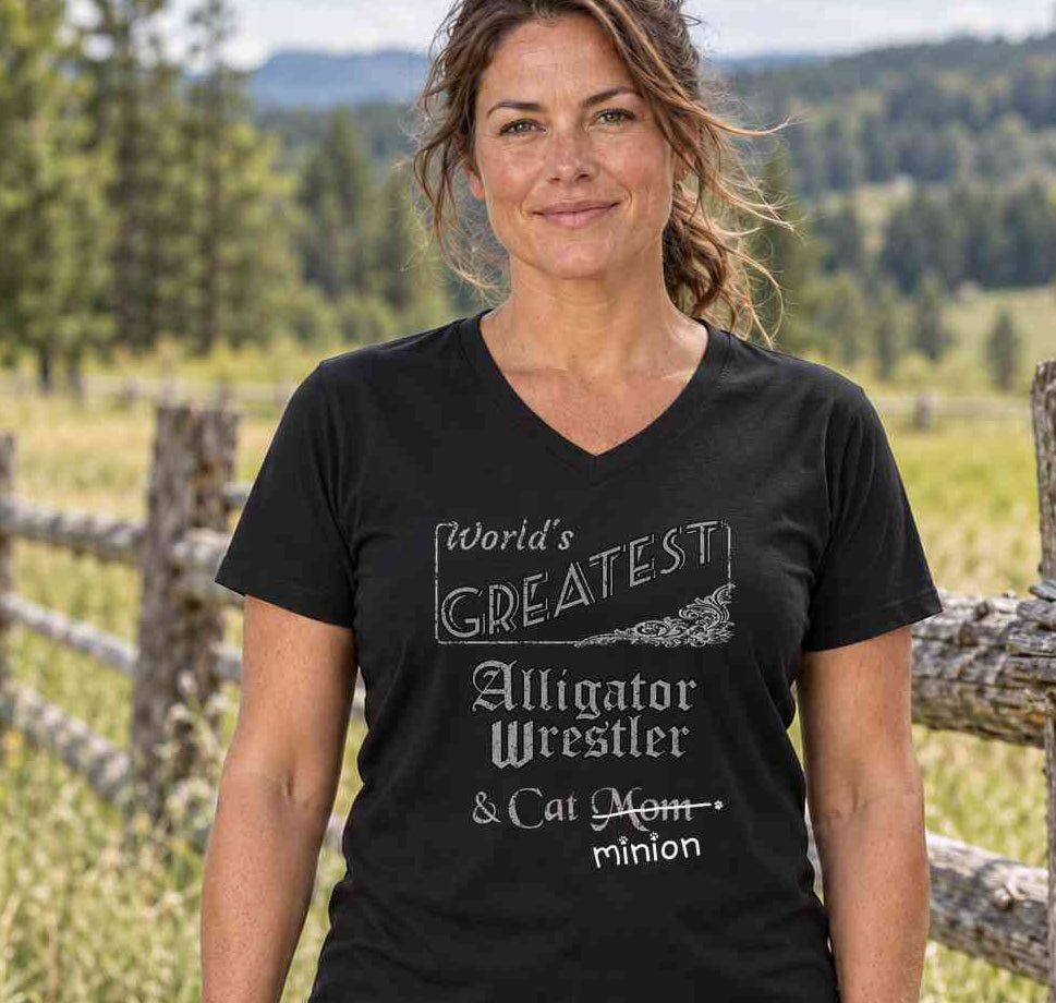 Woman standing beside a wooden fence in the countryside, wearing a black V-neck T-shirt reading “World’s Greatest Alligator Wrestler & Cat Mom.” “Mom” is crossed out, with “minion” handwritten beneath it and surrounded by cat paw prints.