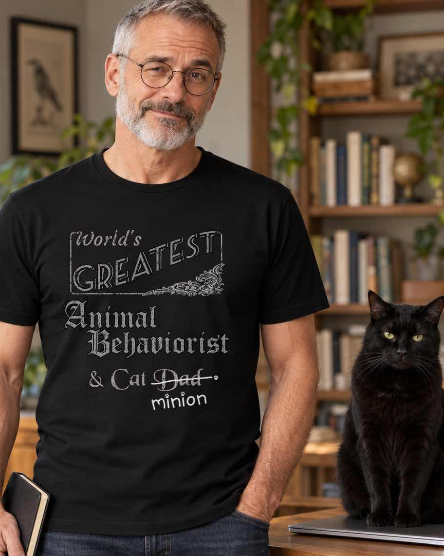 Man holding a notebook beside a cat on a desk, wearing a black crew-neck t-shirt that reads, “World’s Greatest Animal Behaviorist & Cat Dad” with “Dad” crossed out and “minion” handwritten below with paw prints.
