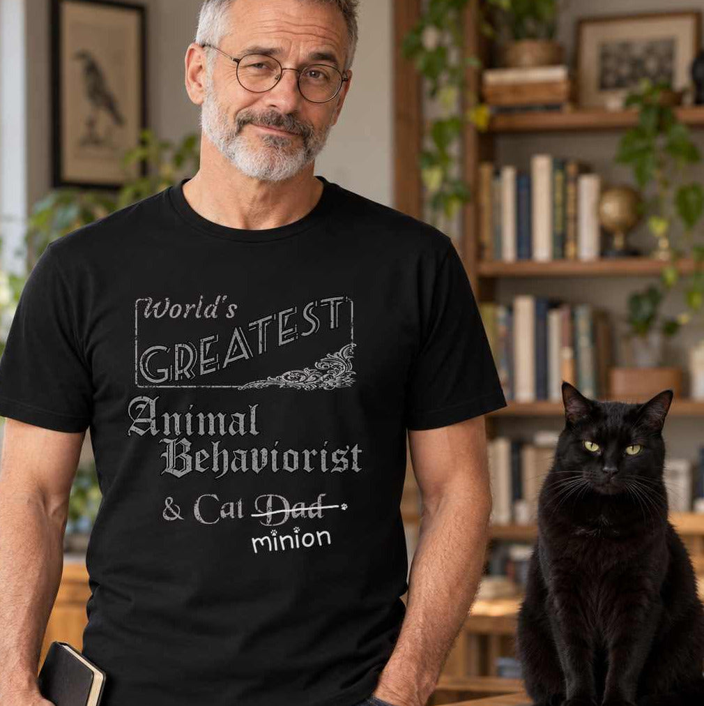 Man holding a notebook beside a cat on a desk, wearing a black crew-neck t-shirt that reads, “World’s Greatest Animal Behaviorist & Cat Dad” with “Dad” crossed out and “minion” handwritten below with paw prints.