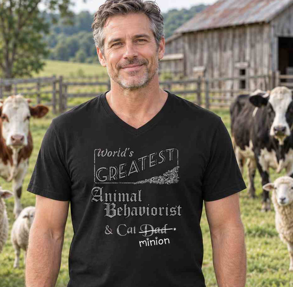 Man standing beside a pasture with cows and sheep, wearing a black V-neck T-shirt reading “World’s Greatest Animal Behaviorist & Cat Dad.” “Dad” is crossed out, with “minion” handwritten beneath it and surrounded by cat paw prints.