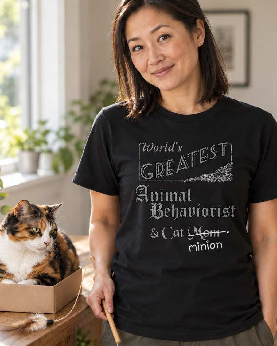 Woman holding a cat toy beside a cat in a home interior, wearing a black crew-neck t-shirt that reads, “World’s Greatest Animal Behaviorist & Cat Mom” with “Mom” crossed out and “minion” handwritten below with paw prints.