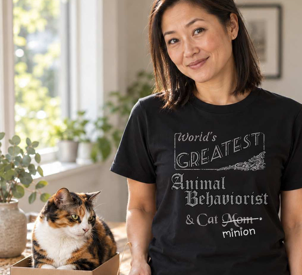 Woman holding a cat toy beside a cat in a home interior, wearing a black crew-neck t-shirt that reads, “World’s Greatest Animal Behaviorist & Cat Mom” with “Mom” crossed out and “minion” handwritten below with paw prints.