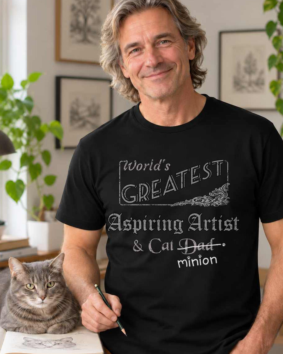 Man standing in an art studio beside a cat, wearing a black crew-neck t-shirt that reads, “World’s Greatest Aspiring Artist & Cat Dad” with “Dad” crossed out and “minion” handwritten below with paw prints.