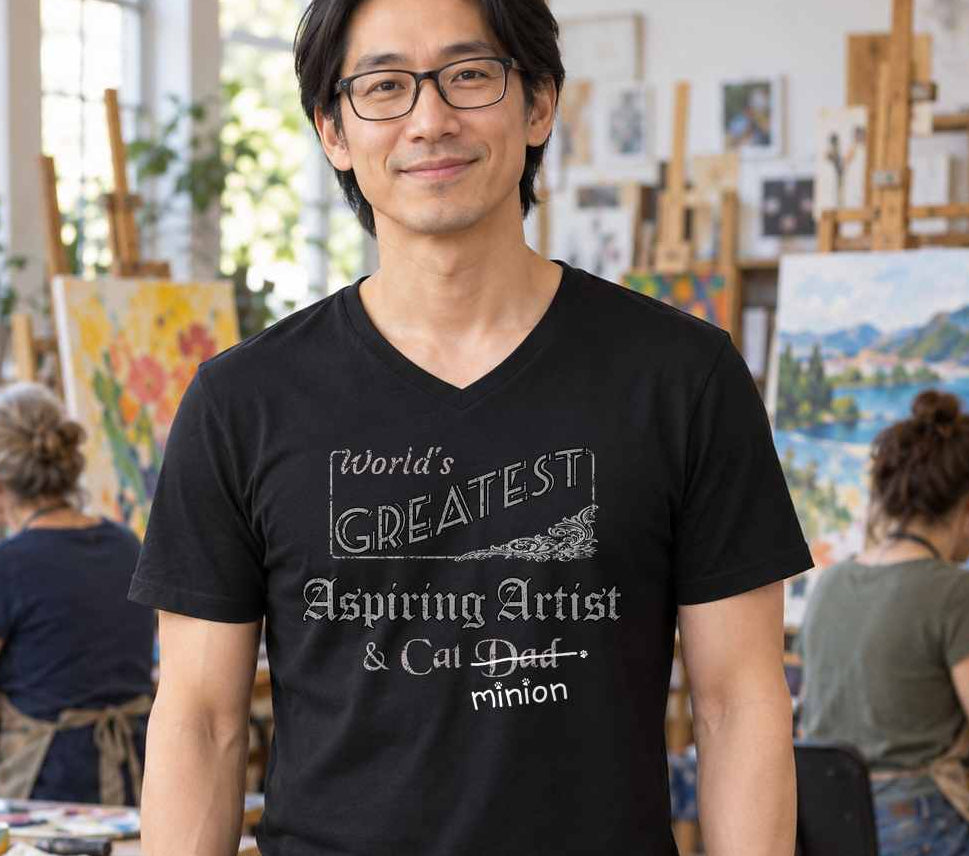 Man standing in an art studio with people painting behind him, wearing a black V-neck T-shirt reading “World’s Greatest Aspiring Artist & Cat Dad.” “Dad” is crossed out, with “minion” handwritten beneath it and surrounded by cat paw prints.