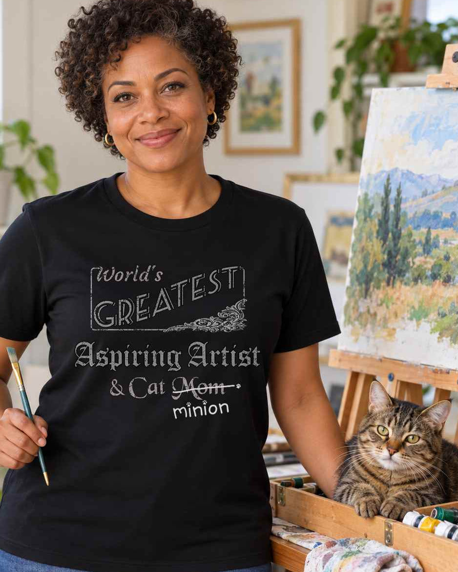 Woman holding a paintbrush in an art studio beside a cat, wearing a black crew-neck t-shirt that reads, “World’s Greatest Aspiring Artist & Cat Mom” with “Mom” crossed out and “minion” handwritten below with paw prints.