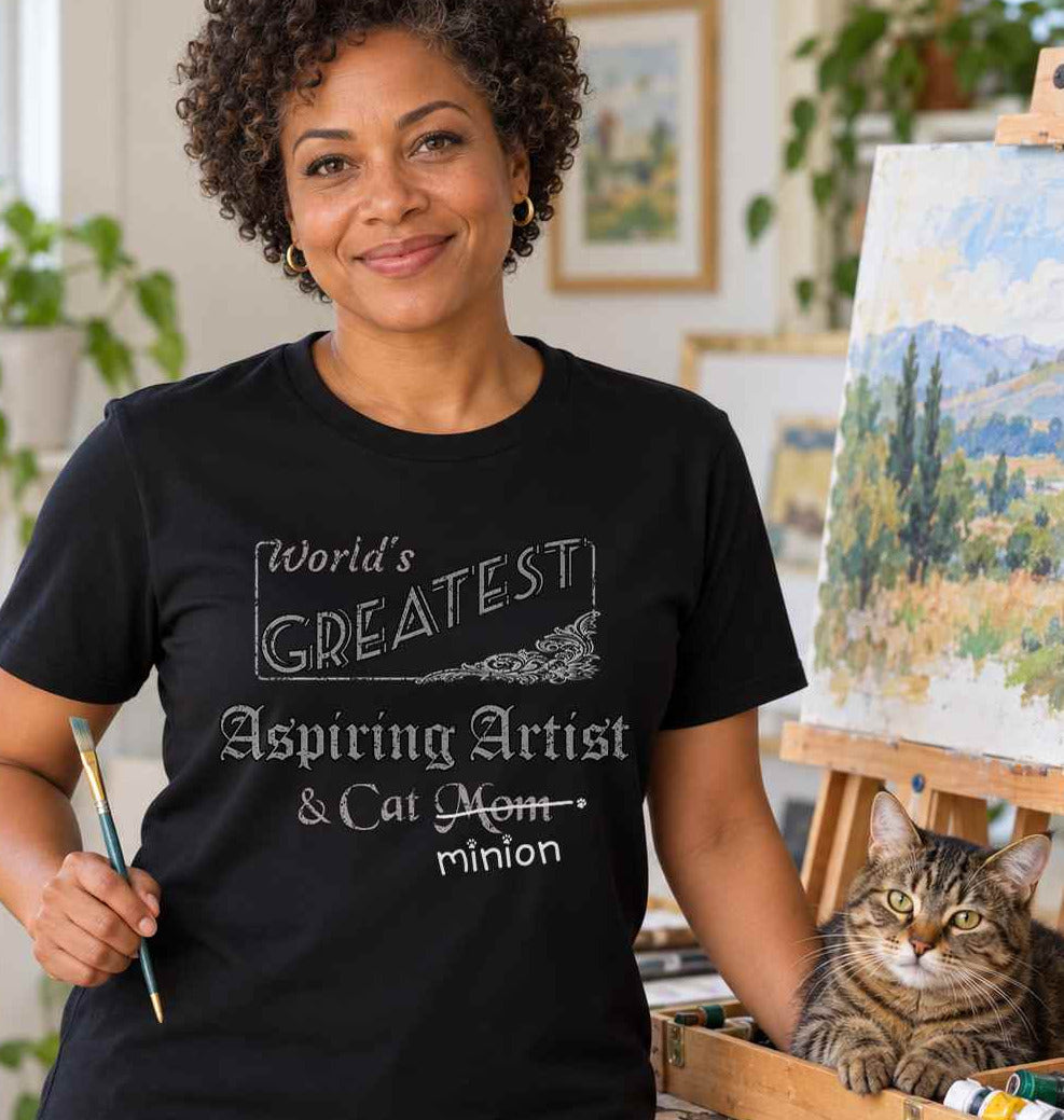 Woman holding a paintbrush in an art studio beside a cat, wearing a black crew-neck t-shirt that reads, “World’s Greatest Aspiring Artist & Cat Mom” with “Mom” crossed out and “minion” handwritten below with paw prints.