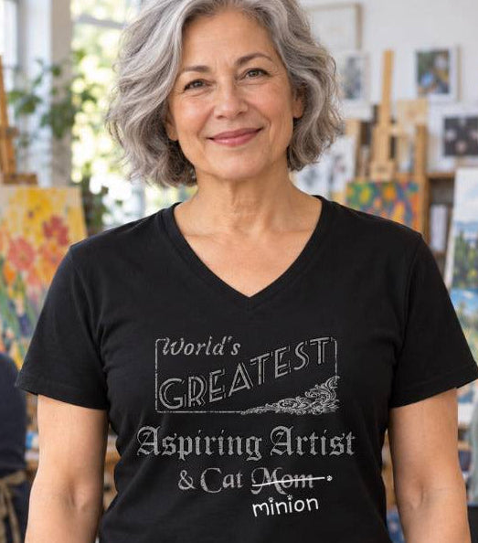 Woman standing in an art studio with people painting behind her, wearing a black V-neck T-shirt reading “World’s Greatest Aspiring Artist & Cat Mom.” “Mom” is crossed out, with “minion” handwritten beneath it and surrounded by cat paw prints.