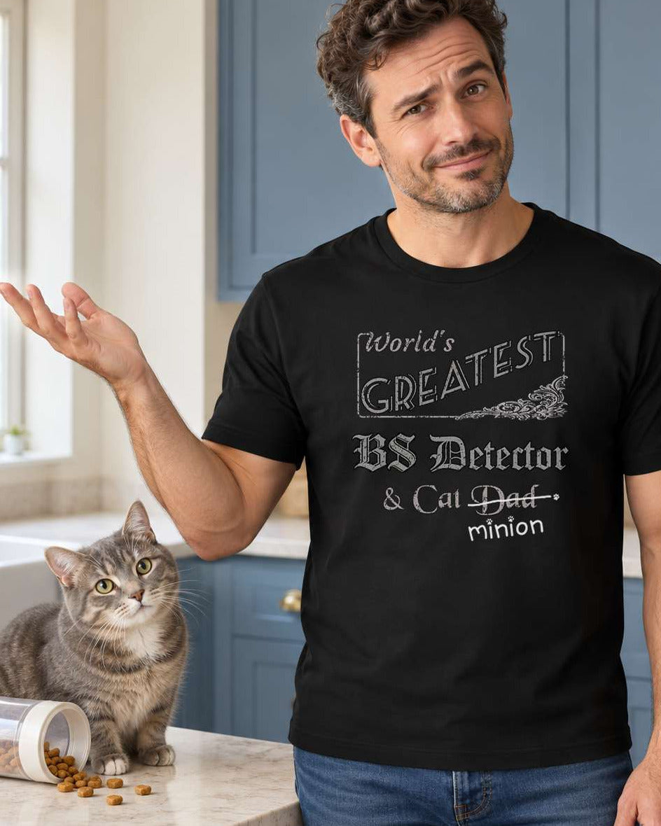 Man gesturing beside a cat in a kitchen, wearing a black crew-neck t-shirt that reads, “World’s Greatest BS Detector & Cat Dad” with “Dad” crossed out and “minion” handwritten below with paw prints.