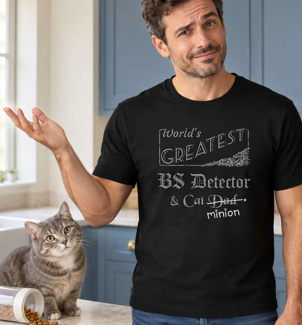 Man gesturing beside a cat in a kitchen, wearing a black crew-neck t-shirt that reads, “World’s Greatest BS Detector & Cat Dad” with “Dad” crossed out and “minion” handwritten below with paw prints.