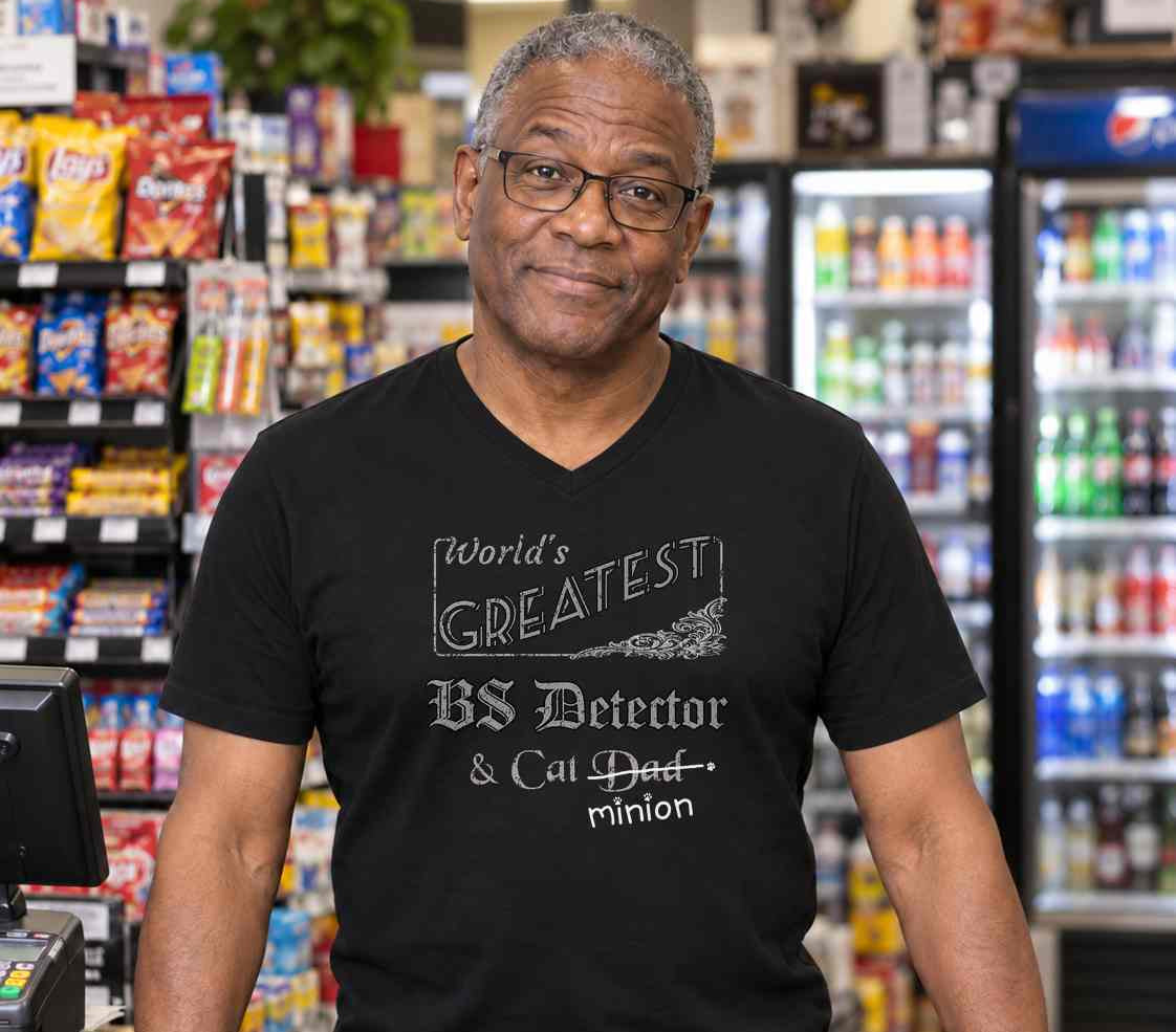 Man standing behind a shop counter, wearing a black V-neck T-shirt reading “World’s Greatest BS Detector & Cat Dad.” “Dad” is crossed out, with “minion” handwritten beneath it and surrounded by cat paw prints.