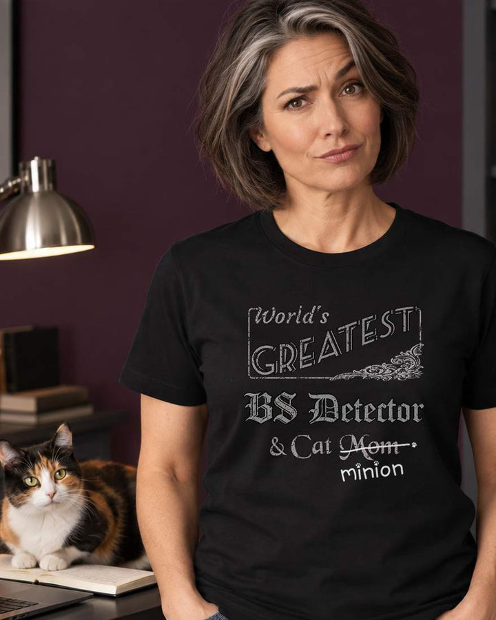 Woman standing beside a cat in a home office, wearing a black crew-neck t-shirt that reads, “World’s Greatest BS Detector & Cat Mom” with “Mom” crossed out and “minion” handwritten below with paw prints.