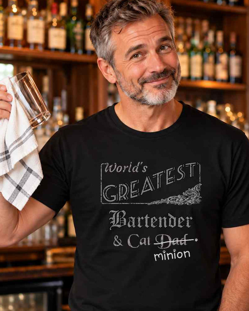 Man holding a bar towel beside a cat on a bar counter, wearing a black crew-neck t-shirt that reads, “World’s Greatest Bartender & Cat Dad” with “Dad” crossed out and “minion” handwritten below with paw prints.