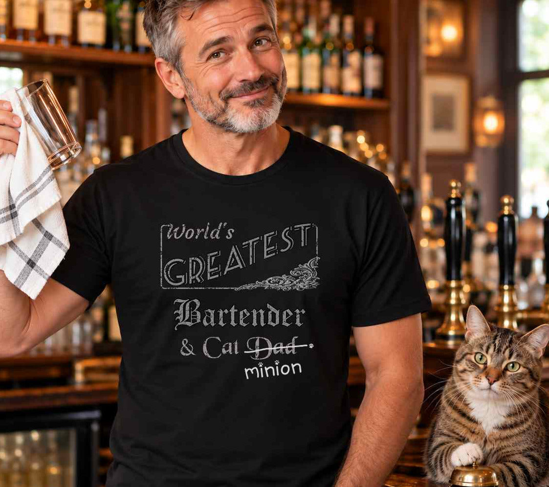 Man holding a bar towel beside a cat on a bar counter, wearing a black crew-neck t-shirt that reads, “World’s Greatest Bartender & Cat Dad” with “Dad” crossed out and “minion” handwritten below with paw prints.