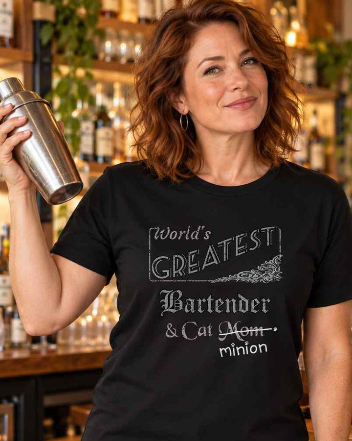 Woman holding a cocktail shaker at a bar beside a cat, wearing a black crew-neck t-shirt that reads, “World’s Greatest Bartender & Cat Mom” with “Mom” crossed out and “minion” handwritten below with paw prints.