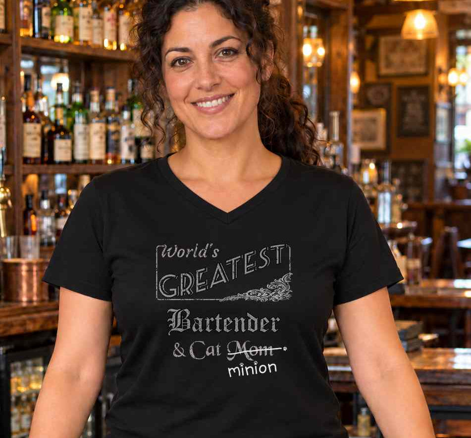 Woman standing behind a pub bar, wearing a black V-neck T-shirt reading “World’s Greatest Bartender & Cat Mom.” “Mom” is crossed out, with “minion” handwritten beneath it and surrounded by cat paw prints.
