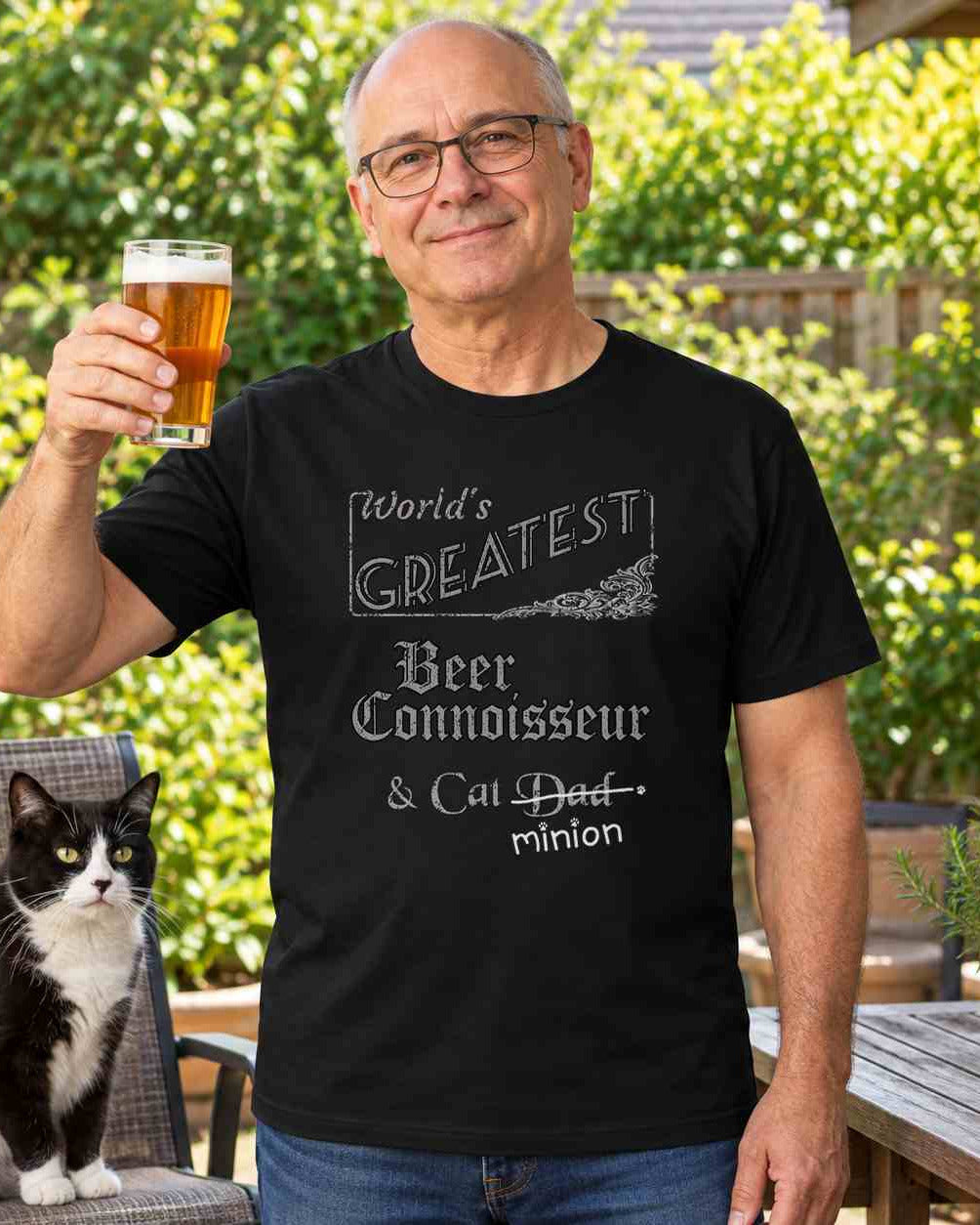 Man raising a glass of beer outdoors beside a cat, wearing a black crew-neck t-shirt that reads, “World’s Greatest Beer Connoisseur & Cat Dad” with “Dad” crossed out and “minion” handwritten below with paw prints.