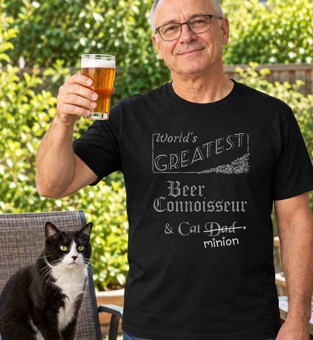 Man raising a glass of beer outdoors beside a cat, wearing a black crew-neck t-shirt that reads, “World’s Greatest Beer Connoisseur & Cat Dad” with “Dad” crossed out and “minion” handwritten below with paw prints.