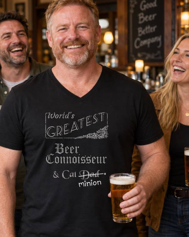 Man holding a pint of beer among friends in a pub, wearing a black V-neck T-shirt reading “World’s Greatest Beer Connoisseur & Cat Dad.” “Dad” is crossed out, with “minion” handwritten beneath it and surrounded by cat paw prints.