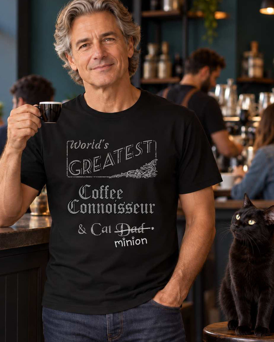 Man holding a coffee cup at a cafe beside a cat, wearing a black crew-neck t-shirt that reads, “World’s Greatest Coffee Connoisseur & Cat Dad” with “Dad” crossed out and “minion” handwritten below with paw prints.