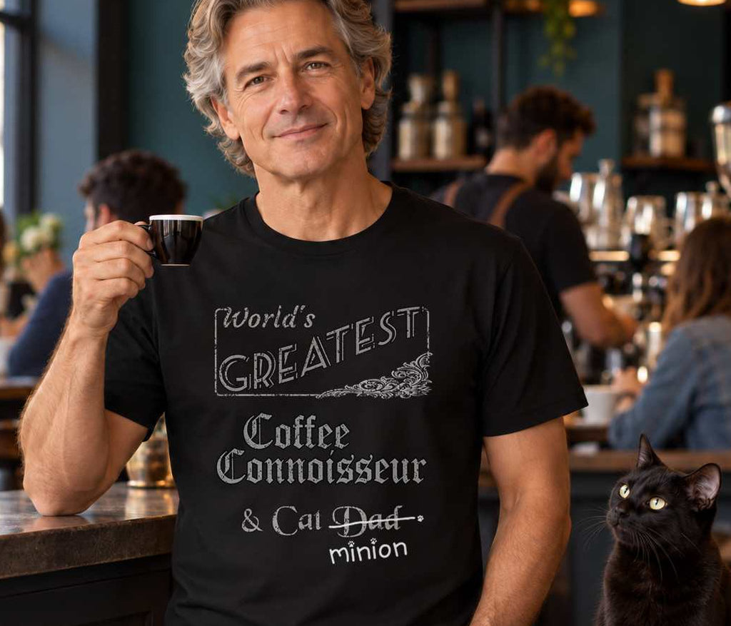 Man holding a coffee cup at a cafe beside a cat, wearing a black crew-neck t-shirt that reads, “World’s Greatest Coffee Connoisseur & Cat Dad” with “Dad” crossed out and “minion” handwritten below with paw prints.