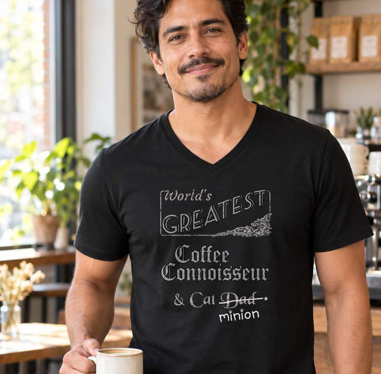 Man holding a mug in a café, wearing a black V-neck T-shirt reading “World’s Greatest Coffee Connoisseur & Cat Dad.” “Dad” is crossed out, with “minion” handwritten beneath it and surrounded by cat paw prints.