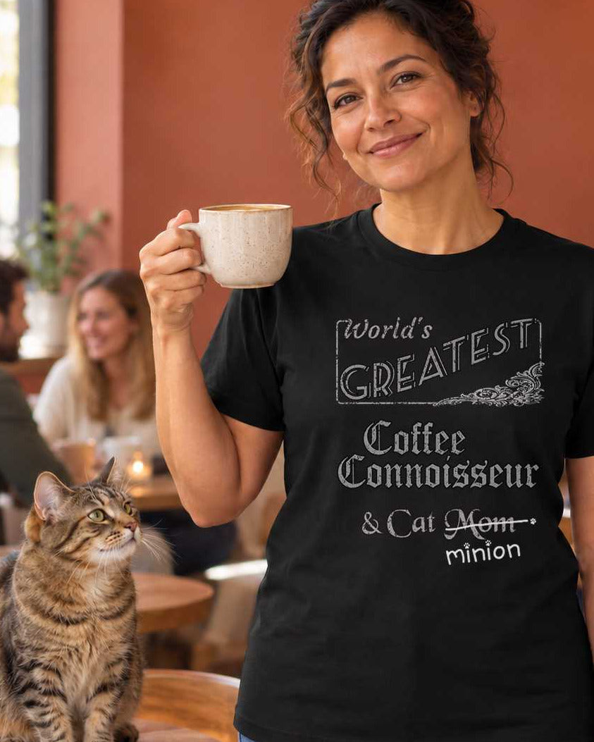 Woman holding a coffee cup at a cafe beside a cat, wearing a black crew-neck t-shirt that reads, “World’s Greatest Coffee Connoisseur & Cat Mom” with “Mom” crossed out and “minion” handwritten below with paw prints.