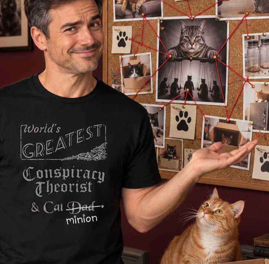 Man gesturing toward a wall of photographs beside a cat, wearing a black crew-neck t-shirt that reads, “World’s Greatest Conspiracy Theorist & Cat Dad” with “Dad” crossed out and “minion” handwritten below with paw prints.