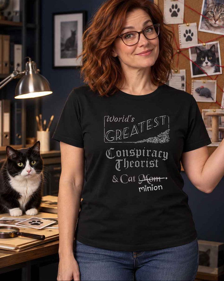 Woman gesturing toward a wall of photographs beside a cat, wearing a black crew-neck t-shirt that reads, “World’s Greatest Conspiracy Theorist & Cat Mom” with “Mom” crossed out and “minion” handwritten below with paw prints.