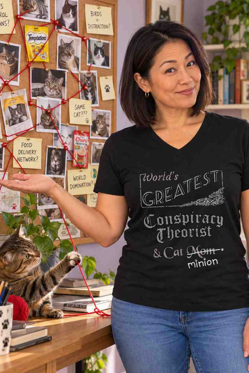 Woman presenting a board of cat photographs connected by red yarn while a cat plays with the yarn, wearing a black V-neck T-shirt reading “World’s Greatest Conspiracy Theorist & Cat Mom.” “Mom” is crossed out, with “minion” handwritten beneath it and surrounded by cat paw prints.
