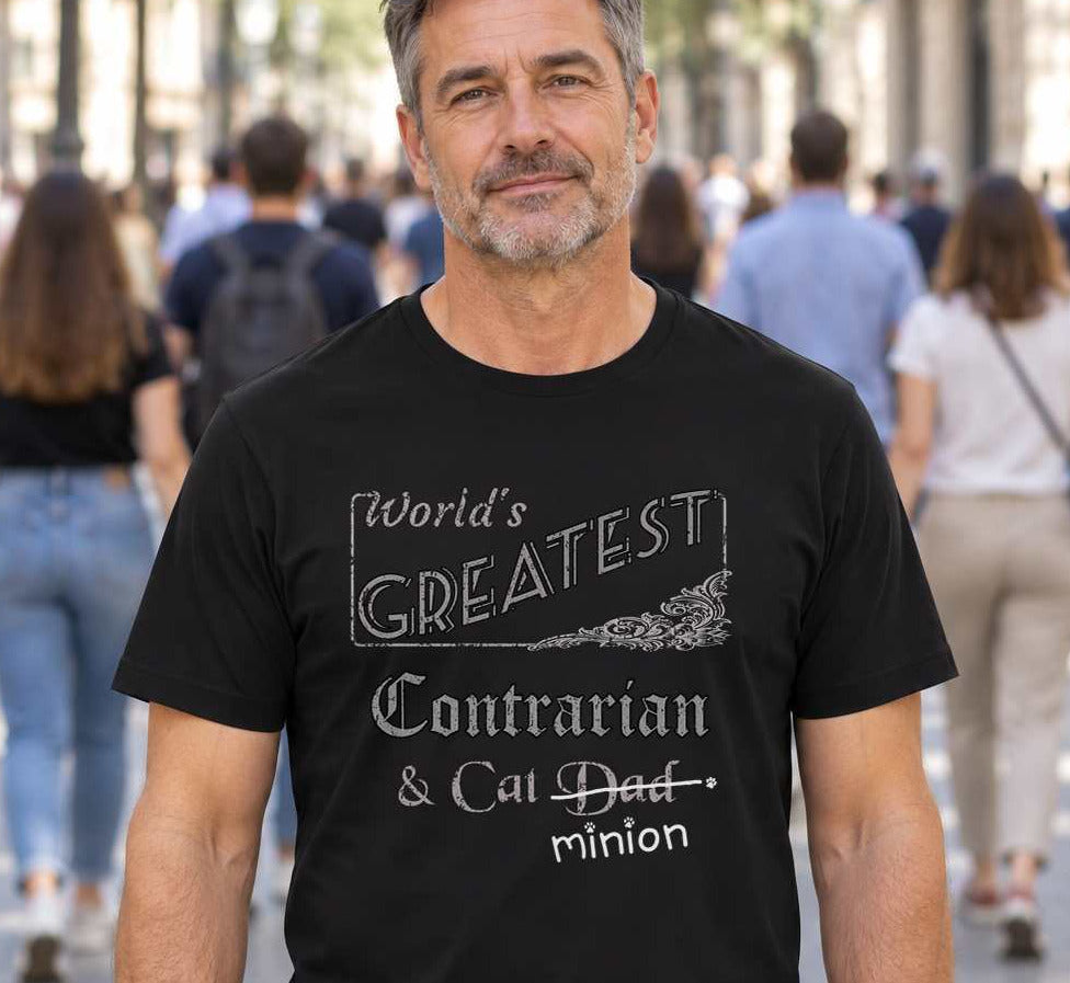 Man facing the camera while a crowd walks away, wearing a black crew-neck t-shirt that reads, “World’s Greatest Contrarian & Cat Dad” with “Dad” crossed out and “minion” handwritten below with paw prints.