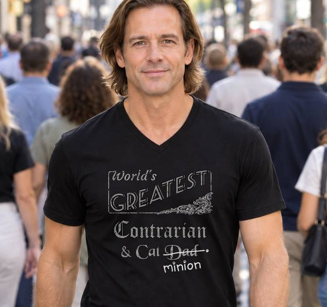 Man standing among pedestrians in a busy street, wearing a black V-neck T-shirt reading “World’s Greatest Contrarian & Cat Dad.” “Dad” is crossed out, with “minion” handwritten beneath it and surrounded by cat paw prints.