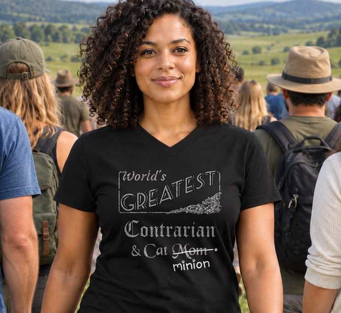 Woman standing outdoors with people behind her, wearing a black V-neck T-shirt reading “World’s Greatest Contrarian & Cat Mom.” “Mom” is crossed out, with “minion” handwritten beneath it and surrounded by cat paw prints.