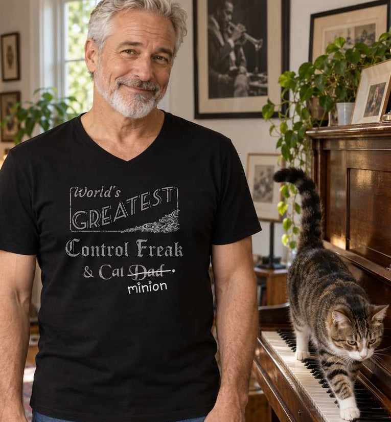 Man standing beside a piano as a cat walks across the keys, wearing a black V-neck T-shirt reading “World’s Greatest Control Freak & Cat Dad.” “Dad” is crossed out, with “minion” handwritten beneath it and surrounded by cat paw prints.